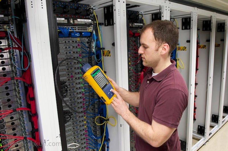 Man standing in front of data server rack, holding a yellow and gray testing device in his hand